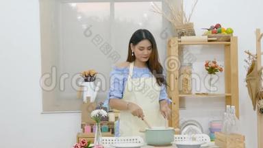 4K. 漂亮的亚洲女人在餐桌后面用勺子搅拌汤，厨房里有锅、菜和厨具